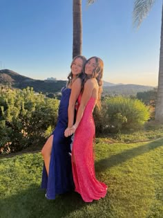 two women in long dresses standing next to each other on the grass with palm trees behind them