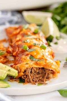 a white plate topped with meat and cheese covered enchilada next to sliced avocado