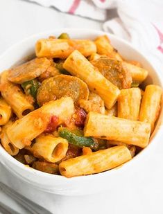 a white bowl filled with pasta covered in sausage and peppers next to a fork on top of a table