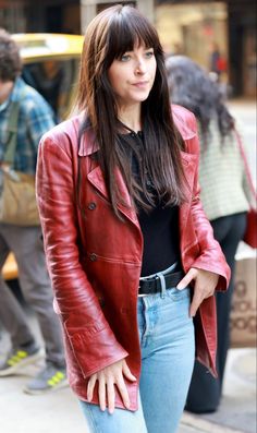 a woman wearing a red leather jacket and jeans standing on the street with her hands in her pockets