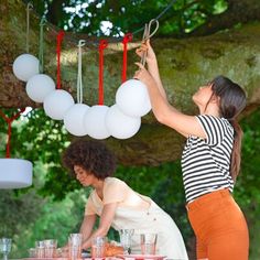 two women hanging balloons from a tree with drinks on the table in front of them