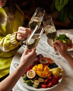 two people toasting with glasses of champagne over a plate of fruit and veggies