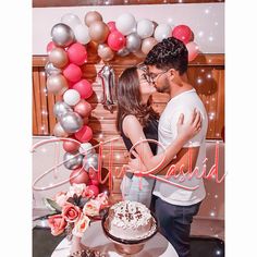 a man and woman kissing in front of a cake with balloons on the wall behind them