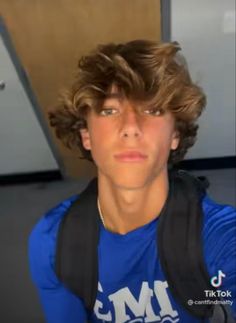 a young man with curly hair wearing a blue shirt and black backpack looking at the camera