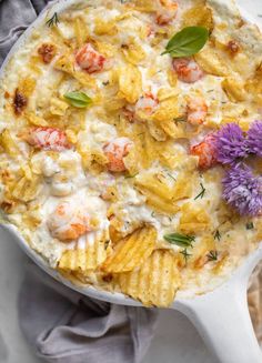 a white casserole dish filled with pasta, shrimp and cream sauce next to a purple flower