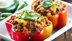 three stuffed peppers on a white plate with green peppers in the background and other vegetables