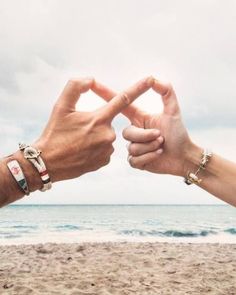 two people making a heart shape with their hands on the beach in front of the ocean