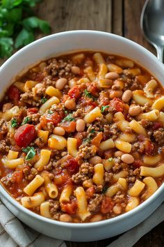 a white bowl filled with pasta and meat soup on top of a table next to a spoon