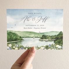 a hand holding up a save the date card with watercolor flowers on it and a lake in the background