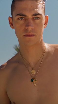 a man with no shirt on standing in the sand and wearing a necklace that has a gold disc attached to it