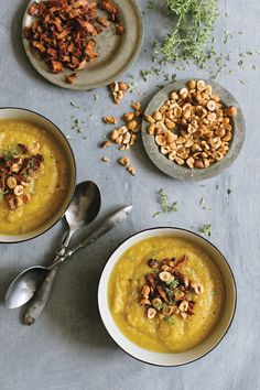 three bowls filled with soup and nuts on top of a gray tablecloth next to spoons