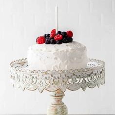 a white cake with berries on top is sitting on a stand and has two candles in the middle