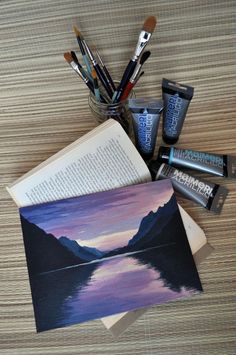 an open book sitting on top of a wooden table next to paint brushes and other art supplies