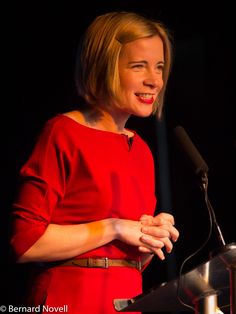 a woman in a red dress standing at a podium with her hands folded and smiling