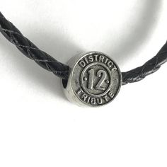 a black leather bracelet with a silver metal button on the clasp that says district 12