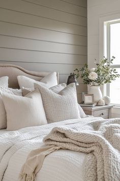 a white bed topped with lots of pillows and blankets next to a vase filled with flowers