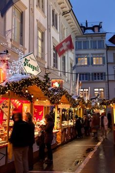 people are shopping at an outdoor market on a city street with christmas lights strung across the buildings