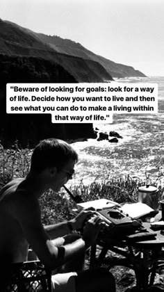 a man sitting at a table with a pen and paper in his hand next to the ocean