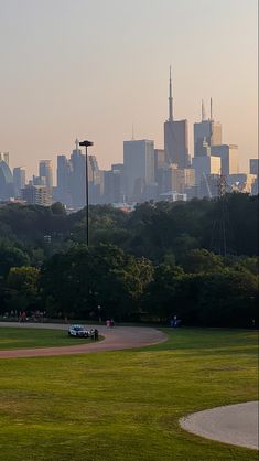 a city skyline is seen in the distance behind a baseball field with cars parked on it