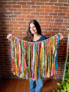 a woman holding up a colorful scarf in front of a brick wall