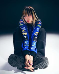 a young woman sitting on the ground with her hands in her pockets, wearing a blue jacket and black pants