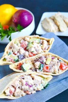 two pita breads filled with chicken salad on top of a blue napkin next to bowls of vegetables