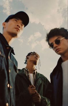 three young men standing next to each other under a cloudy blue sky with clouds in the background