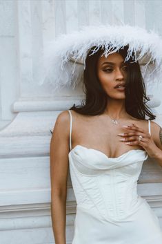 a woman in a white dress and feathered hat poses for a photo with her hands on her chest