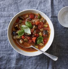 a white bowl filled with vegetable soup next to a spoon on top of a blue cloth