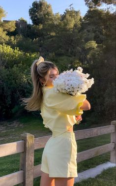 a woman in yellow dress holding white flowers by wooden fence with grass and trees behind her