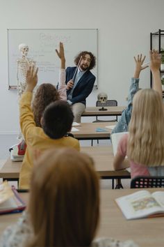 a group of children sitting at desks in front of a whiteboard with their hands up