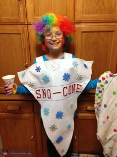 a child wearing a snow cone costume standing in front of a wooden cabinet holding a cup