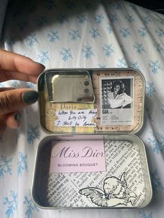 an open tin with some pictures in it on top of a sheet covered bed next to a person's hand