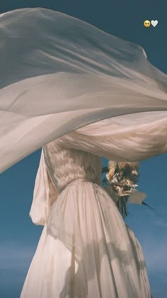 a woman in a white dress is holding her veil over her head and looking up at the sky