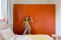 a woman standing in front of an orange wall next to a white bed and chair