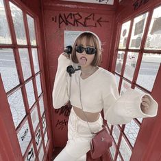 a woman in white clothes and sunglasses talking on a phone while standing next to a red telephone booth