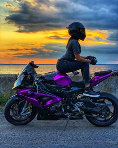 a woman sitting on top of a purple motorcycle next to the ocean at sunset or dawn