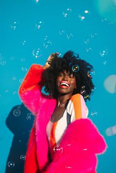 a woman with curly hair wearing a bright pink coat and posing in front of bubbles