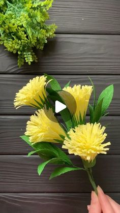 someone is holding yellow flowers in front of a wooden wall with green leaves on it