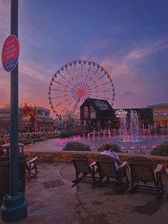 people are sitting on benches in front of a water fountain and a ferris wheel at dusk