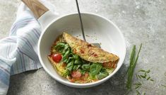 an omelet with spinach and tomatoes in a white bowl next to a wooden spoon