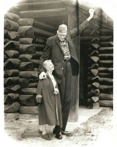 an old photo of a man and woman standing in front of a pile of logs