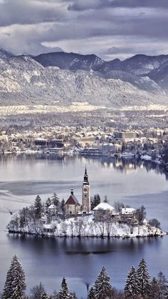 an island in the middle of a lake surrounded by trees and snow covered mountains with a church on it