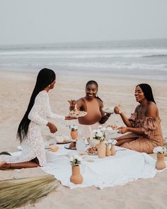 three women sitting on the beach having a picnic with food and drinks in front of them