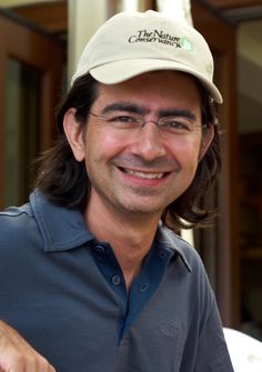 a man with long hair wearing a hat and glasses smiles at the camera while standing in front of a building