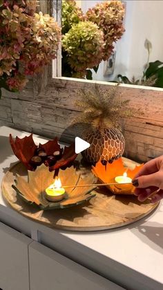 a person lighting candles on a tray with leaves and flowers in front of a mirror