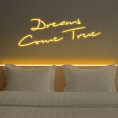 a bed with white sheets and pillows under a neon sign that reads dreams come true