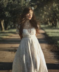 a woman in a white dress is standing on a dirt road with trees behind her