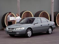 two people standing next to a gray car in front of barrels with wine on them