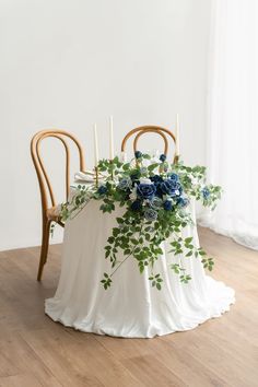 a white table topped with blue flowers and greenery next to two wooden chairs on top of a hard wood floor